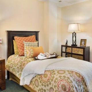 A cozy bedroom interior featuring a neatly made bed with decorative pillows, a lamp on a dresser, and warm lighting.