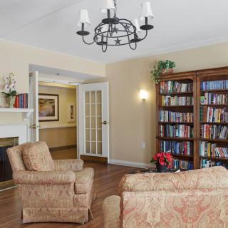 A cozy living room featuring comfortable chairs, a large bookshelf filled with books, a classic chandelier, and a fireplace, all set on a polished wood floor.