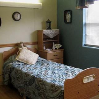 A cozy bedroom featuring a single bed with a quilted blanket, a small shelving unit with decorative items, and a window letting in natural light.