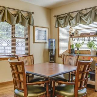 A cozy dining area with a wooden table and chairs, surrounded by windows with curtains and adorned with indoor plants.
