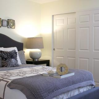 A neatly arranged bedroom with a bed, side table, and lamp, featuring decorative pillows and a closet in the background.