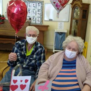 An elderly couple wearing masks, holding heart-shaped balloons and gift bags, sitting indoors celebrating Valentine's Day.