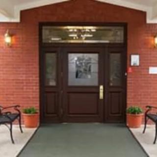 A brick building entrance with a dark wooden door, flanked by potted plants and benches.
