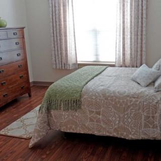 A cozy bedroom with a neatly made bed, a wooden dresser, and soft lighting from the window.