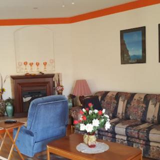A cozy living room featuring a patterned sofa, a blue armchair, and a wooden coffee table with a vase of flowers. The room is decorated with framed pictures and has a fireplace.
