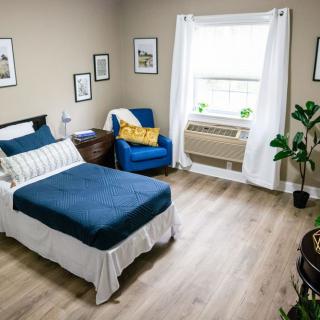 A minimalist bedroom with a single bed, blue chair, and window adorned with white curtains.