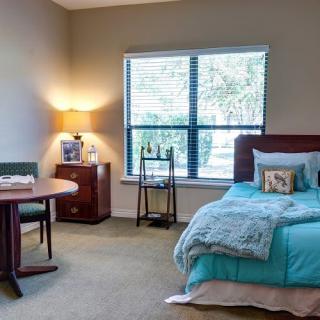 A cozy bedroom with a neatly made bed, a small table and chair, and a lamp by the window.