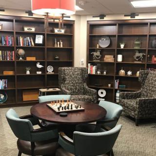 A cozy library corner featuring bookshelves, a chess set on a round table, and comfortable seating.