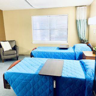 A well-lit hospital room featuring two patient beds with blue bedding, an armchair, and a window allowing natural light.