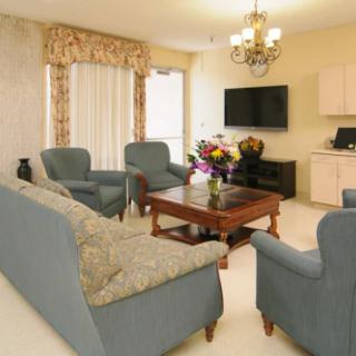 A cozy living room featuring a sofa, armchairs, and a wooden coffee table with flowers. A television is mounted on the wall, and a chandelier adds elegance to the space.