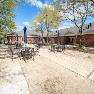A sunny outdoor patio area with tables and chairs, surrounded by trees and a brick building.