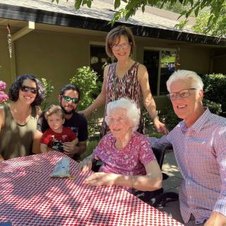 A multi-generational family gathering outdoors, seated around a checkered tablecloth.