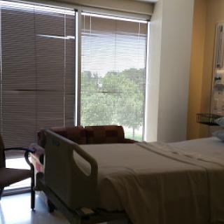 A view of a hospital patient room featuring a bed, chair, and windows with blinds.