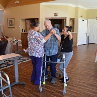 Caregivers assist an elderly man with a walker, providing support and encouragement.