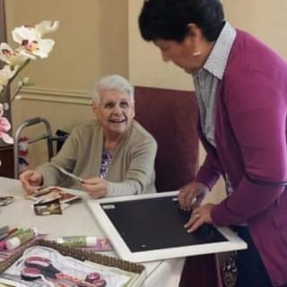 An elderly woman smiles while interacting with a caregiver at a table filled with photos.