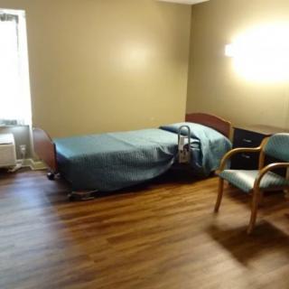A simple, minimalistic room with a bed, chair, and window on a hardwood floor.
