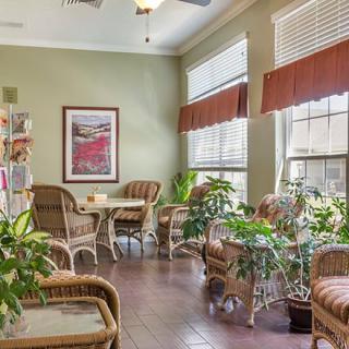 A cozy living room with wicker furniture and lush plants, bathed in natural light from large windows.
