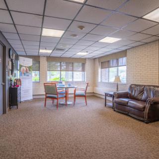 A spacious office area featuring a cozy sofa and a meeting table with chairs, illuminated by natural light from large windows.