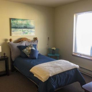 A cozy bedroom with a single bed featuring blue bedding and decorative pillows, complemented by a nightstand and a large window.