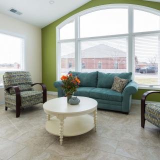 A bright living room with a teal sofa, patterned chairs, and a round coffee table in front of large windows.