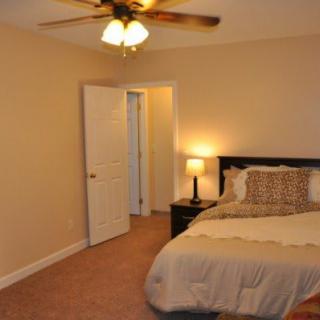 A cozy bedroom featuring a neatly made bed, a nightstand with a lamp, beige walls, and a ceiling fan.