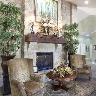 A cozy living room with a stone fireplace, surrounded by armchairs and decorated with indoor plants and artwork.