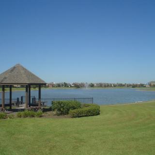 A tranquil lakeside view with a gazebo and lush green grass under a clear blue sky.