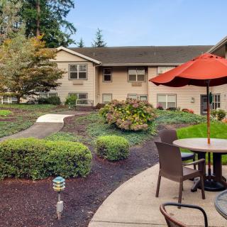 A peaceful garden patio with seating and an umbrella, surrounded by manicured landscaping and adjacent to a residential building.
