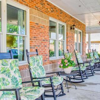 A row of rocking chairs with patterned cushions on a porch of a brick building, accompanied by potted plants.