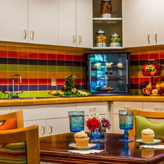 A vibrant kitchen features colorful striped tiles, a cozy dining area with cupcakes and glassware, accented by fresh flowers and decorative elements.
