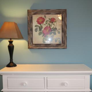 A cozy interior scene featuring a lamp, framed flower artwork, and a white dresser against a blue wall.