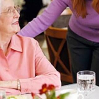 An elderly woman sitting at a dining table being assisted by another person.