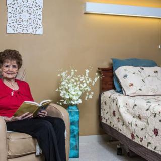 An elderly woman sitting in a comfortable chair next to a cozy bed, reading a book.