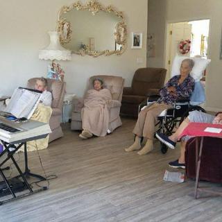 A group of elderly individuals sitting in a cozy living room, some wrapped in blankets and one in a wheelchair, gathered near a piano.