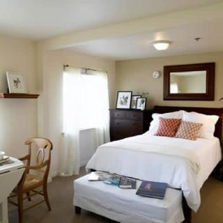 A cozy bedroom with a neatly made bed, mirror, and natural light from the window.