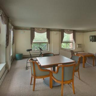 A spacious living room with tables, chairs, and exercise equipment by the windows.