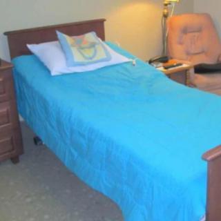 A cozy bedroom with a single bed featuring a blue bedspread, accompanied by a wooden nightstand, lamp, and chair.