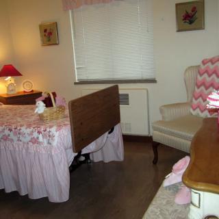 A cozy bedroom with pink decor, featuring a bed, armchair, lamp, and window with curtains.