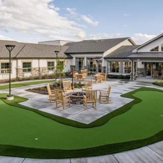 An outdoor patio featuring a fire pit, surrounded by a seating area and artificial grass with a building in the background.