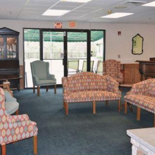 A lounge area with various chairs and sofas arranged on a carpeted floor, featuring large windows in the background.
