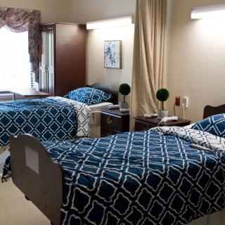A well-organized room with two twin beds featuring blue patterned bedspreads, suitable for a nursing home or hospital setting.