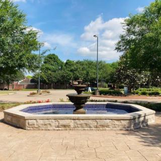 A stone fountain in a landscaped outdoor setting with trees and a clear blue sky.