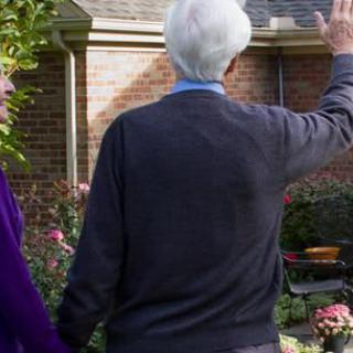 An elderly couple waving in their garden outside a brick house.