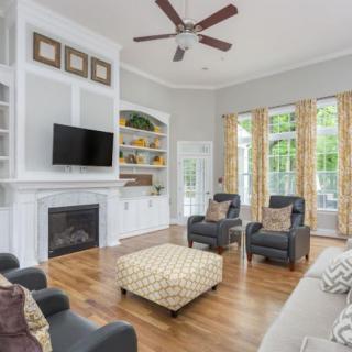 A spacious and elegantly designed living room featuring a fireplace, television, comfortable seating, and large windows with drapes.