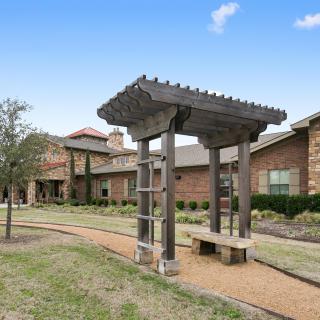 A garden area featuring a wooden pergola along a gravel pathway with a building in the background.