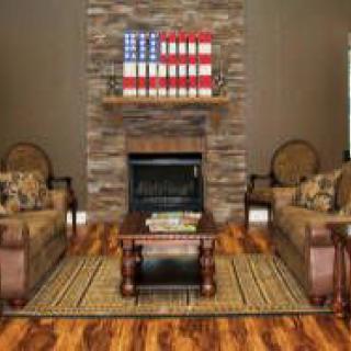 A cozy living room features a central stone fireplace, flanked by two plush couches, with an American flag decoration above and a wooden coffee table on a woven rug.