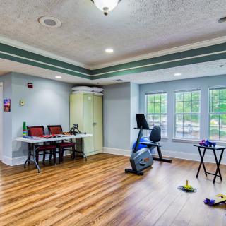 A spacious exercise room with a stationary bike, desk, and chairs on wood flooring.