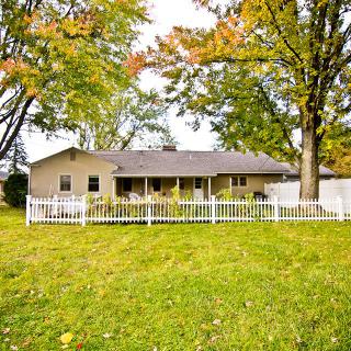 A single-story house with a white fence and a spacious yard surrounded by trees in autumn foliage.