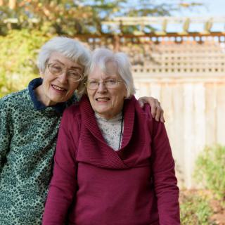 Two elderly women smiling and standing close together outdoors.