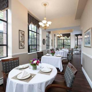 An elegant dining room with neatly arranged tables and chairs, featuring tablecloths, floral centerpieces, large windows, and a chandelier.